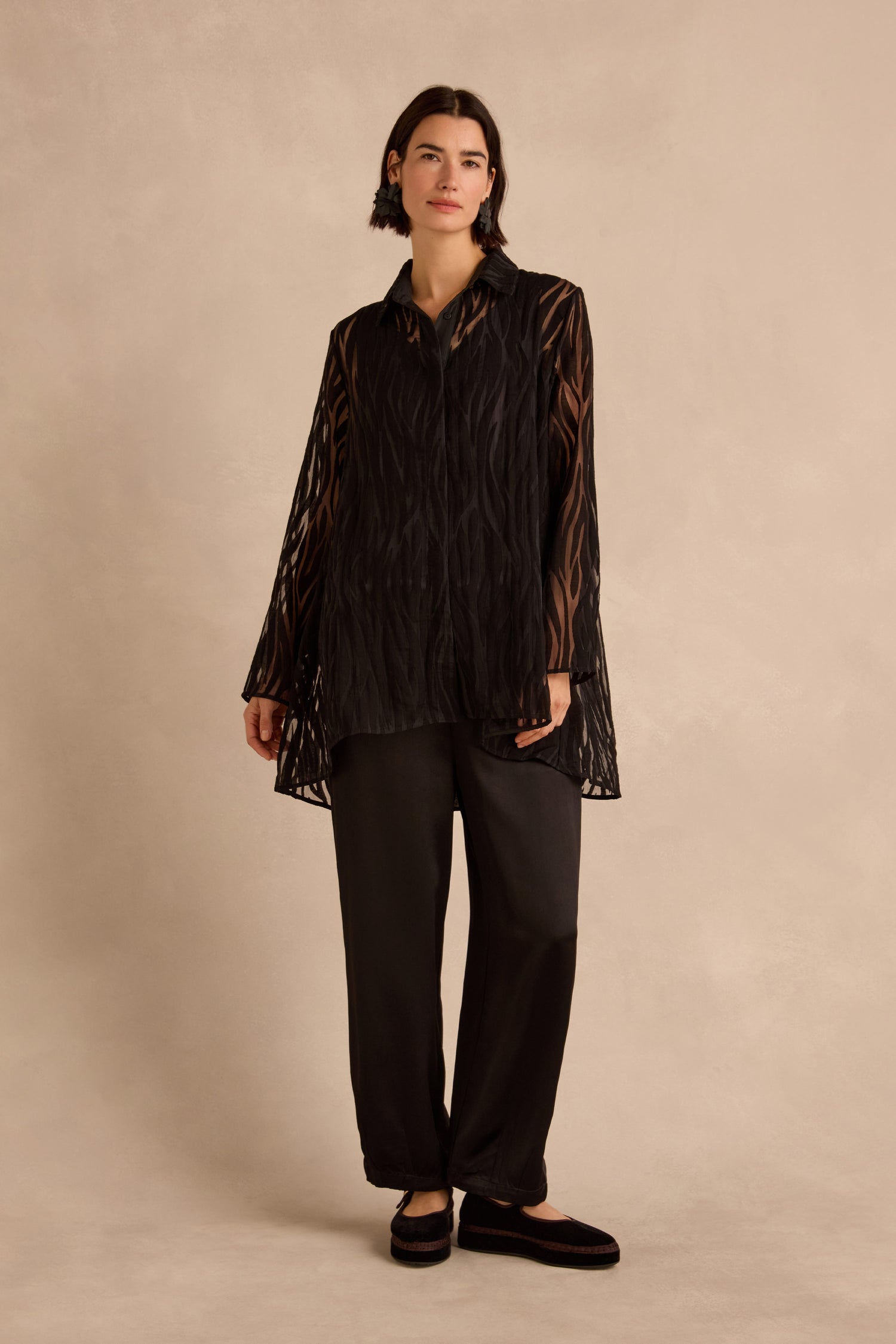A person stands against a beige background wearing the Wave Cut Out Shirt—a sheer, viscose-blend tunic—layered over a black top and paired with loose black pants and black shoes.