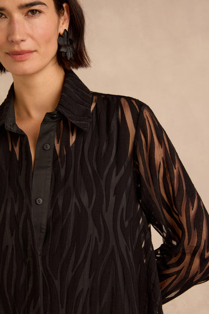 Woman wearing the Wave Cut Out Long Shirt—a sheer black button-up crafted from a viscose blend with a wavy stripe pattern—and black flower earrings, photographed from the shoulders up.