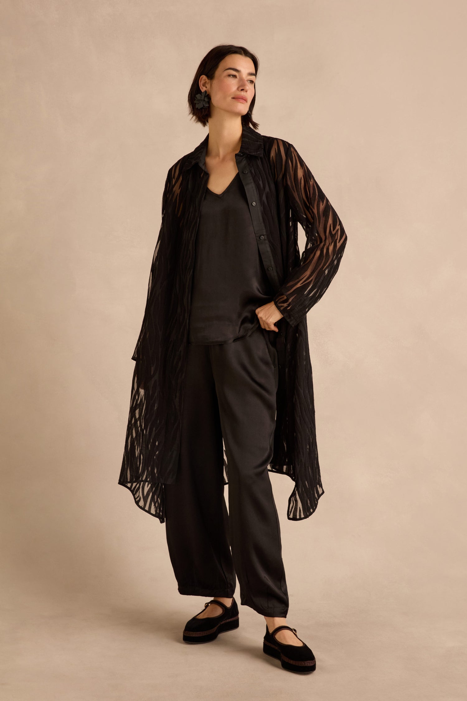 A woman stands wearing the Wave Cut Out Long Shirt, a sheer black long shirt in a viscose blend, styled over a dark top and pants with black flat shoes, posing against a neutral background.