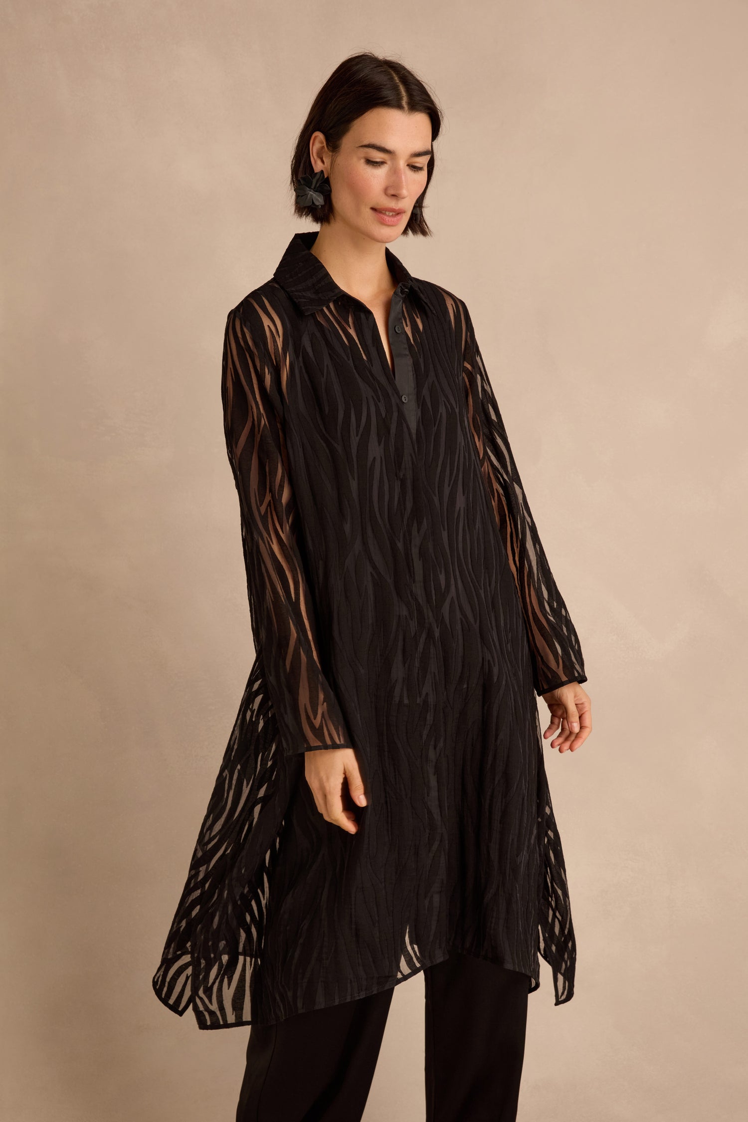 A woman models the Wave Cut Out Long Shirt—a sheer, viscose-blend design featuring a collared neckline, long sleeves, and a subtle wavy pattern with wave cut out detailing—styled over black pants against a neutral background.