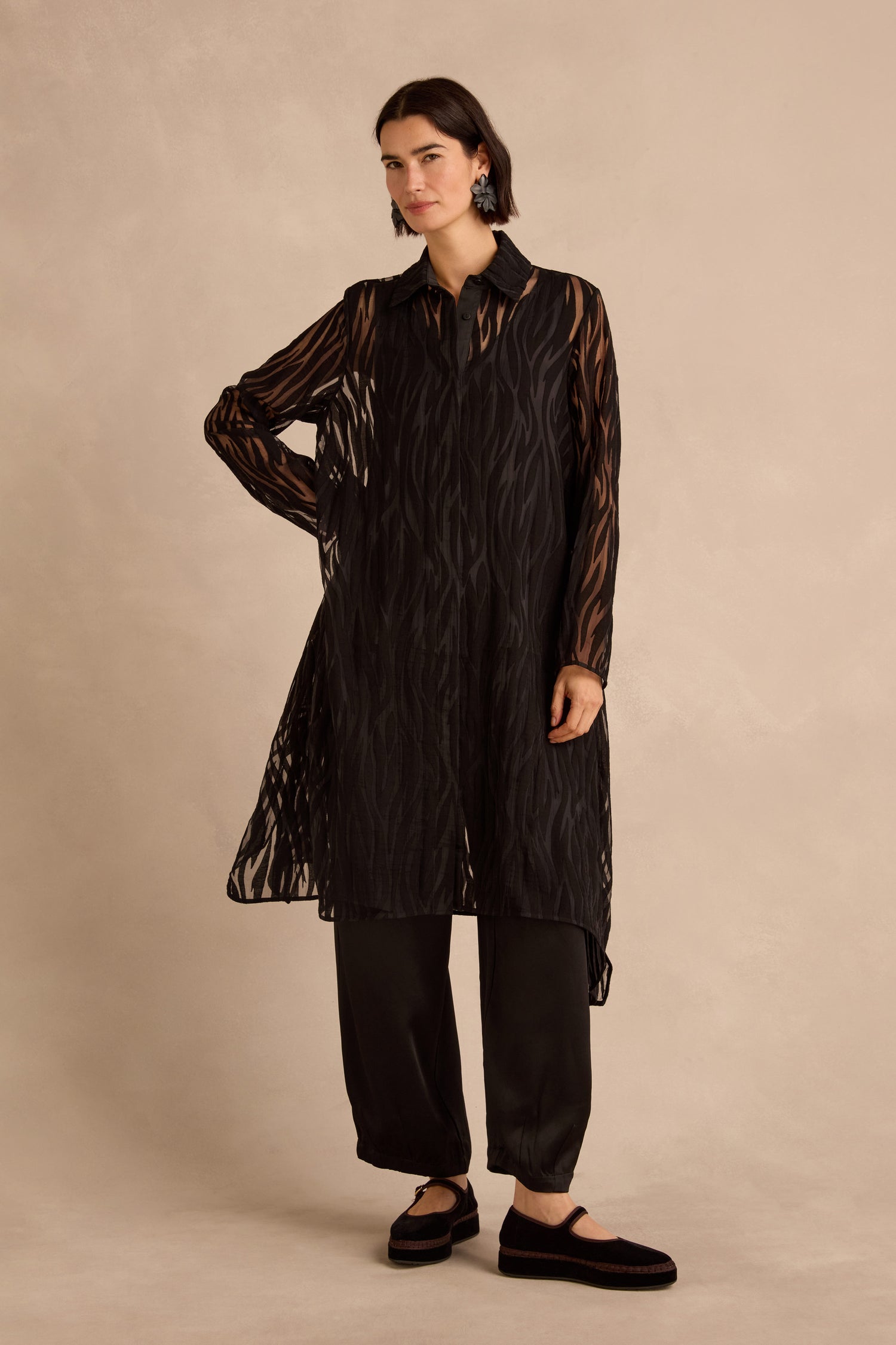 A person stands against a beige background wearing the Wave Cut Out Long Shirt, a sheer piece crafted from a viscose blend with a wavy pattern, styled over black pants and flat black shoes.