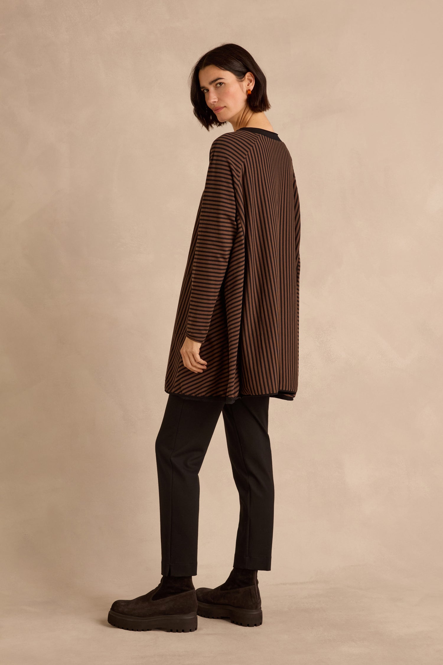 A person is standing sideways against a plain beige background, wearing the Stripe Jersey Wide Neck Tunic—featuring loose brown and black stripes—paired with black pants and black platform shoes.