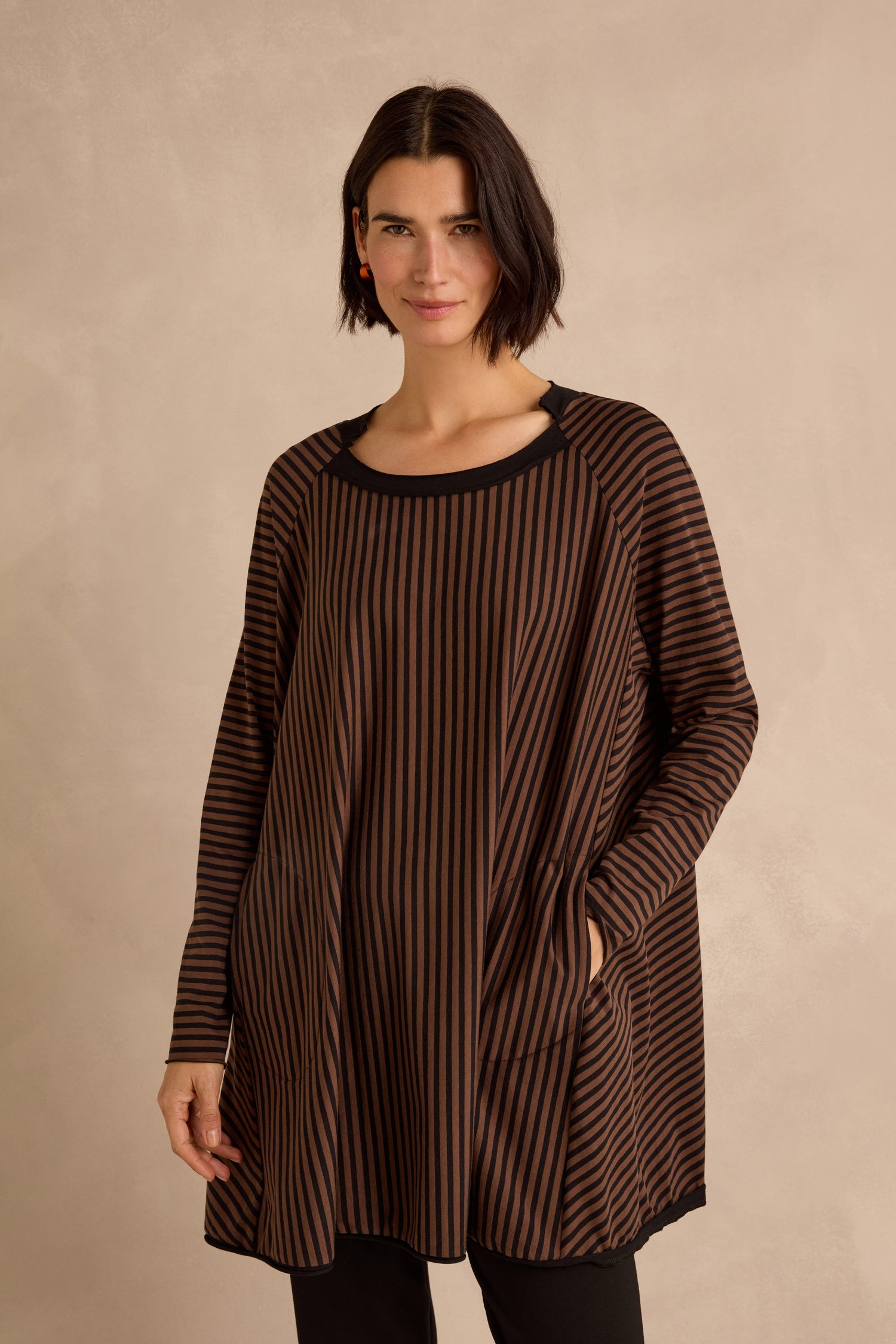 A woman stands against a neutral background wearing the Stripe Jersey Wide Neck Tunic, featuring black and brown vertical stripes and crafted from comfortable cotton stretch jersey with a loose fit.