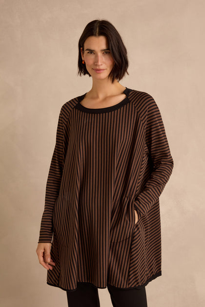 A woman stands with her hands in the pockets of the Stripe Jersey Wide Neck Tunic, featuring a loose fit with bold black stripes, a wide notched neckline, and a striped hem. Crafted from soft cotton stretch jersey for effortless comfort, she poses against a plain beige background.
