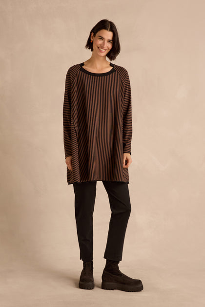 A woman stands against a beige backdrop wearing the Stripe Jersey Wide Neck Tunic, featuring a loose silhouette, wide notched neckline, and striped accents. She pairs it with black pants and shoes, her dark hair and neutral expression effortlessly highlighting the comfortable cotton stretch jersey fabric.