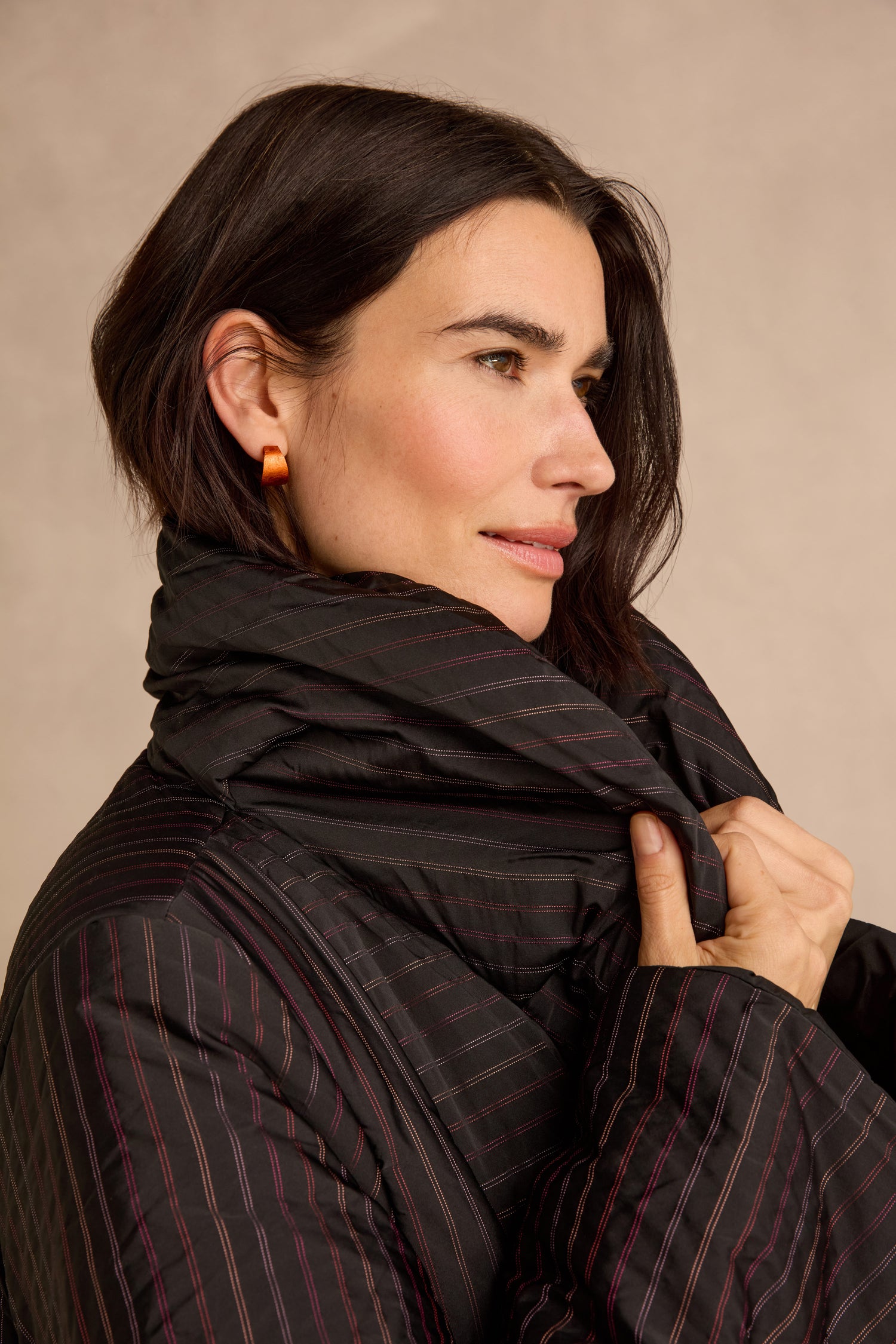 A woman with short dark hair wears orange earrings and the Striped Puffer Coat from Luukaa outerwear, holding the collar up around her neck as she looks to the side against a neutral background.