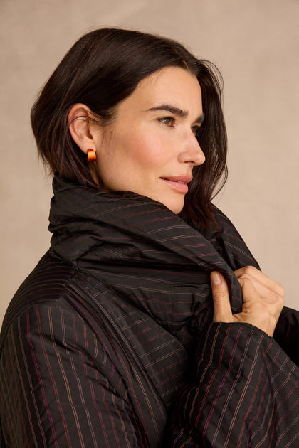 A woman with short dark hair wears orange earrings and the Striped Puffer Coat from Luukaa outerwear, holding the collar up around her neck as she looks to the side against a neutral background.