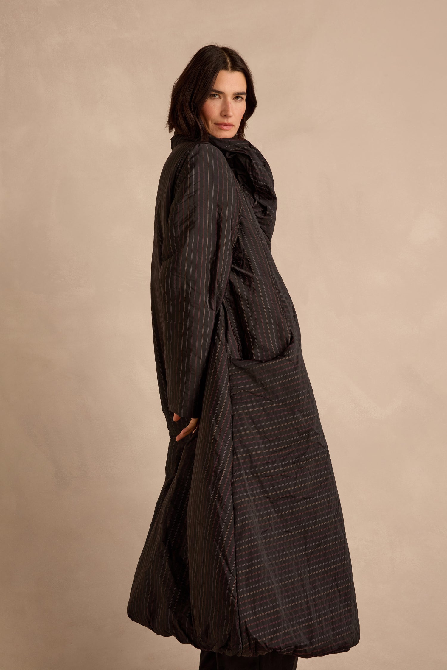 A person with short dark hair wears a long, oversized Striped Puffer Coat and stands against a plain beige background.