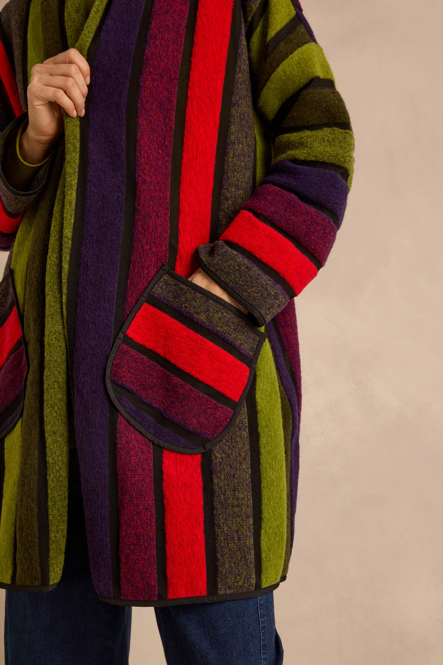 A person wearing the Sheer Stripe Jacket, a vibrant long-sleeved wool-blend piece featuring vertical red, green, purple, and black stripes and large front pockets. Only the jacket and torso are shown.
