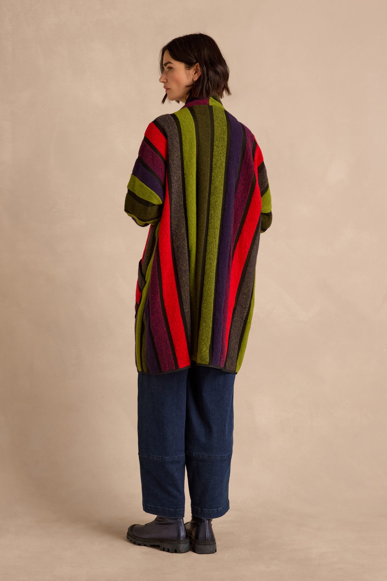 A person stands sideways against a beige background, wearing the Sheer Stripe Jacket featuring multicoloured red, green, purple, and brown stripes, paired with wide-leg blue jeans and black boots.