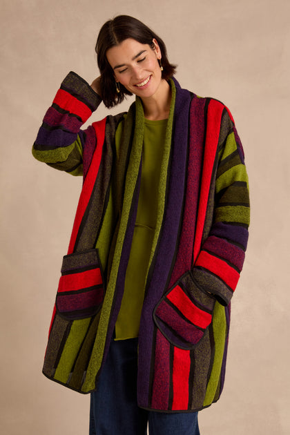 A woman is wearing the Sheer Stripe Jacket—an oversized piece featuring multicoloured stripes in red, purple, green, and brown—layered over a green shirt. She stands with one hand raised near her head and smiles.