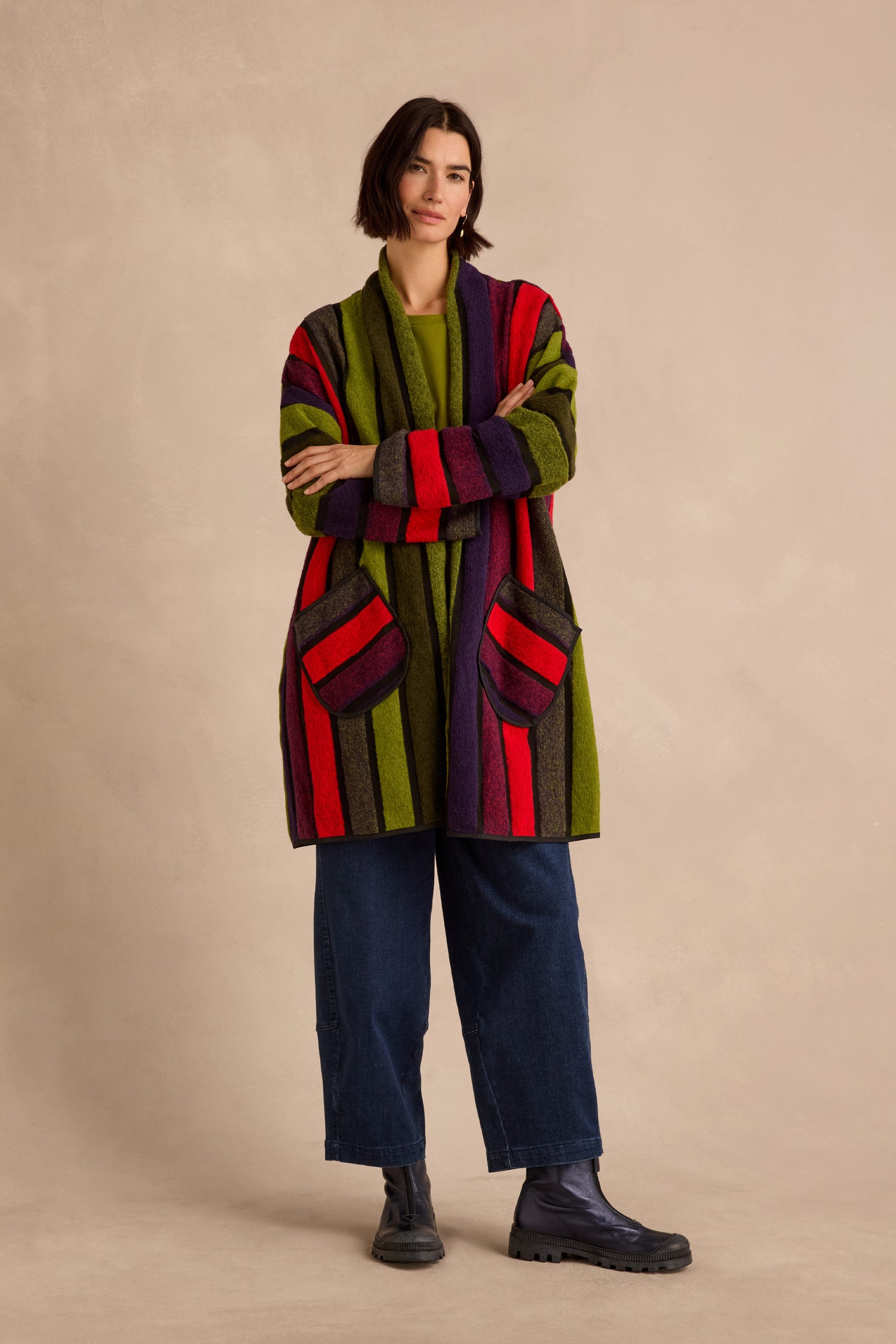 A person stands with arms crossed in front of a plain beige background, wearing the Sheer Stripe Jacket—a long, multicoloured piece featuring green, red, and purple stripes—paired with dark wide-leg jeans and black boots.