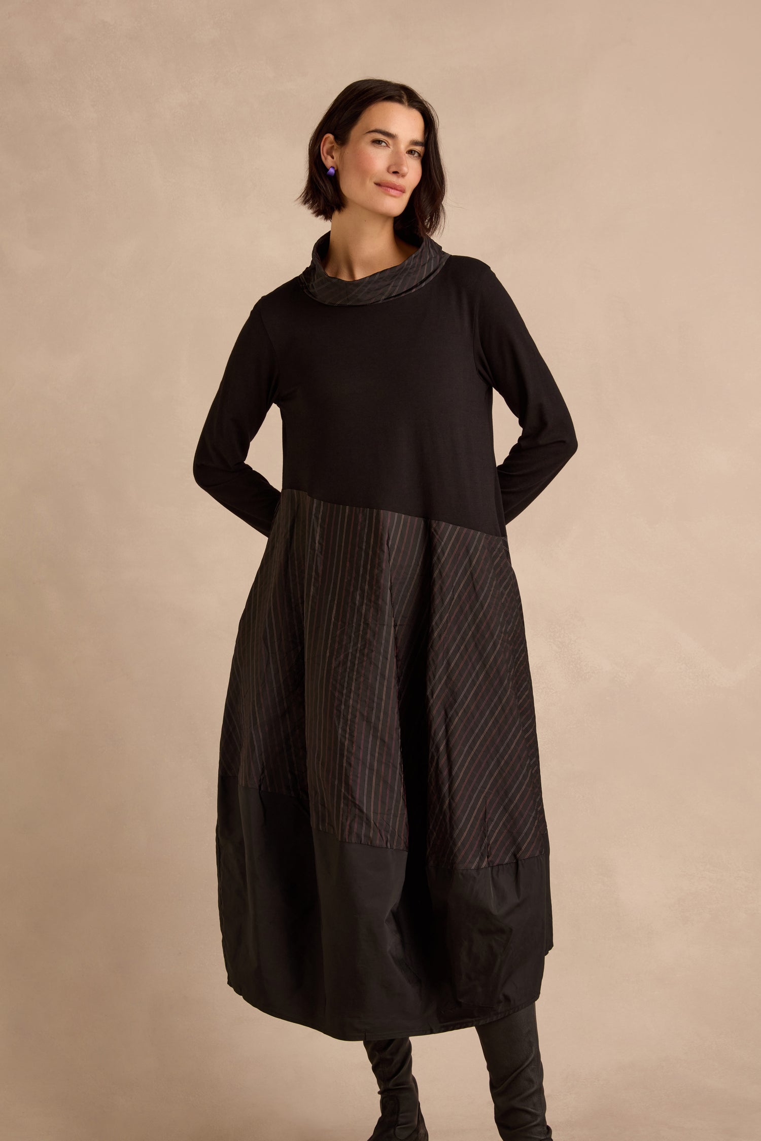A woman stands against a beige background wearing the Stripe Jersey Taffeta Bubble Dress, which features long sleeves, a cowl neck, a textured skirt, and is paired with black boots.
