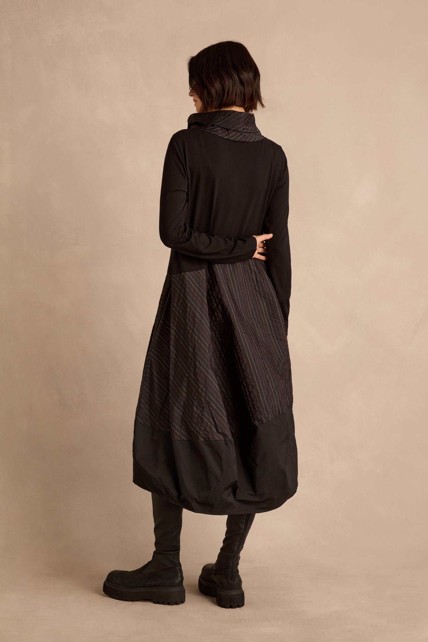 A person stands with their back turned, wearing the Stripe Jersey Taffeta Bubble Dress featuring long sleeves and a textured skirt, paired with a black scarf and black boots against a neutral background.
