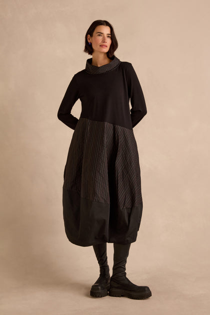 A woman wearing the Stripe Jersey Taffeta Bubble Dress—a long-sleeved, brown and black striped dress with pockets—paired with black leggings and black boots, stands against a neutral beige background.
