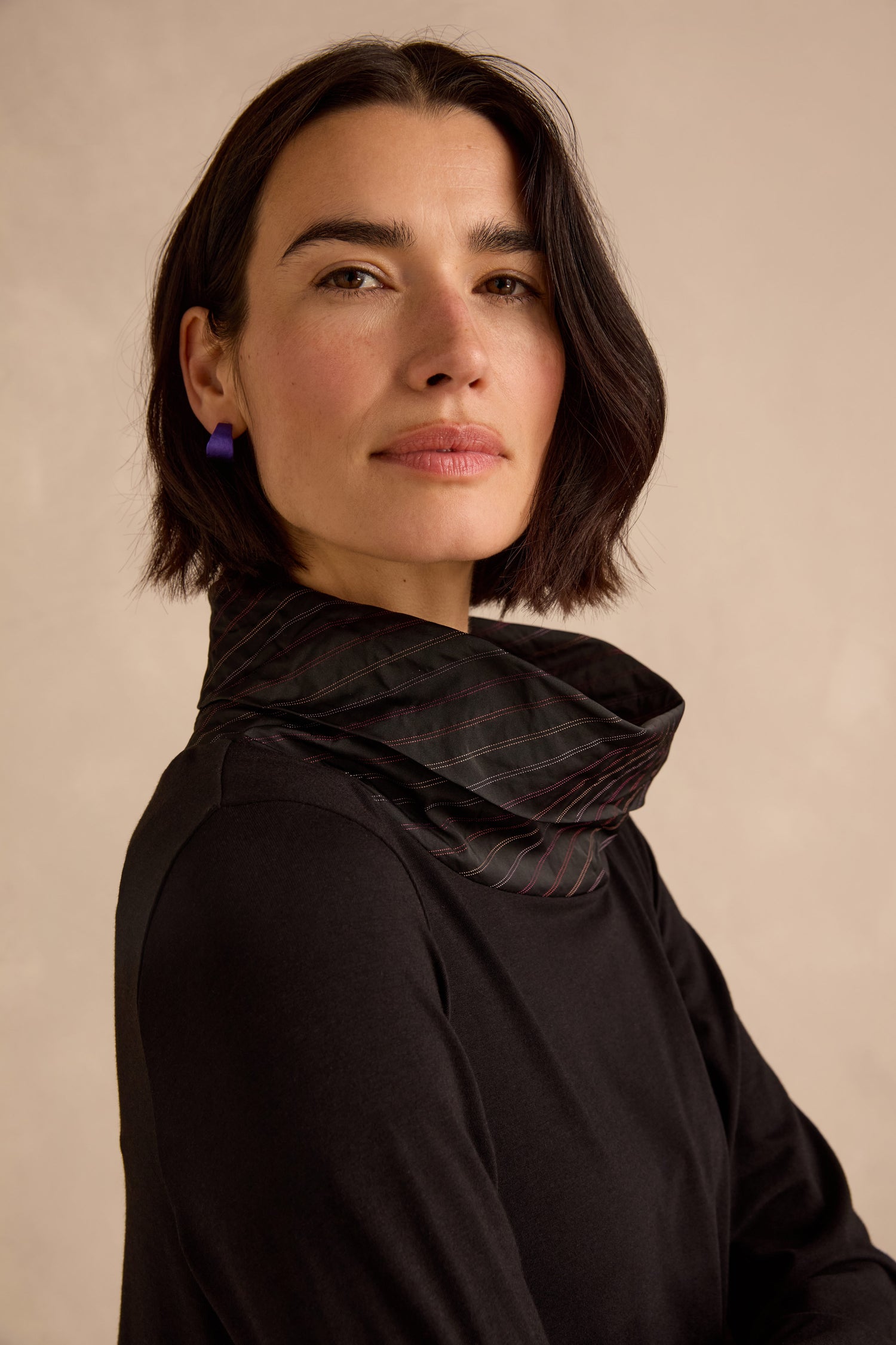 Woman with short dark hair wearing a black top, a patterned black scarf, and purple earrings, standing against a plain beige background, looking slightly to the side. Her modern style is reminiscent of the effortless charm found in the Stripe Jersey Taffeta Bubble Dress.
