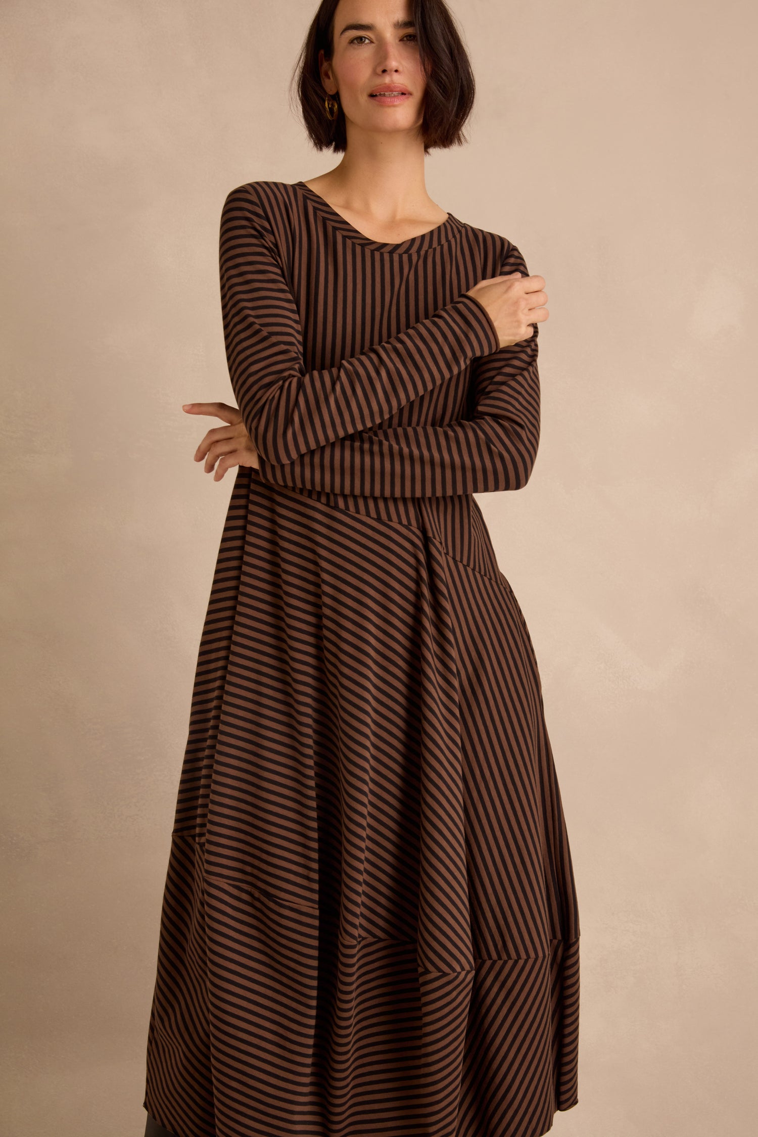 A woman stands against a beige background, wearing the Draped Stripe Jersey Dress—a long-sleeve brown and black striped jersey dress featuring an asymmetrical hem.