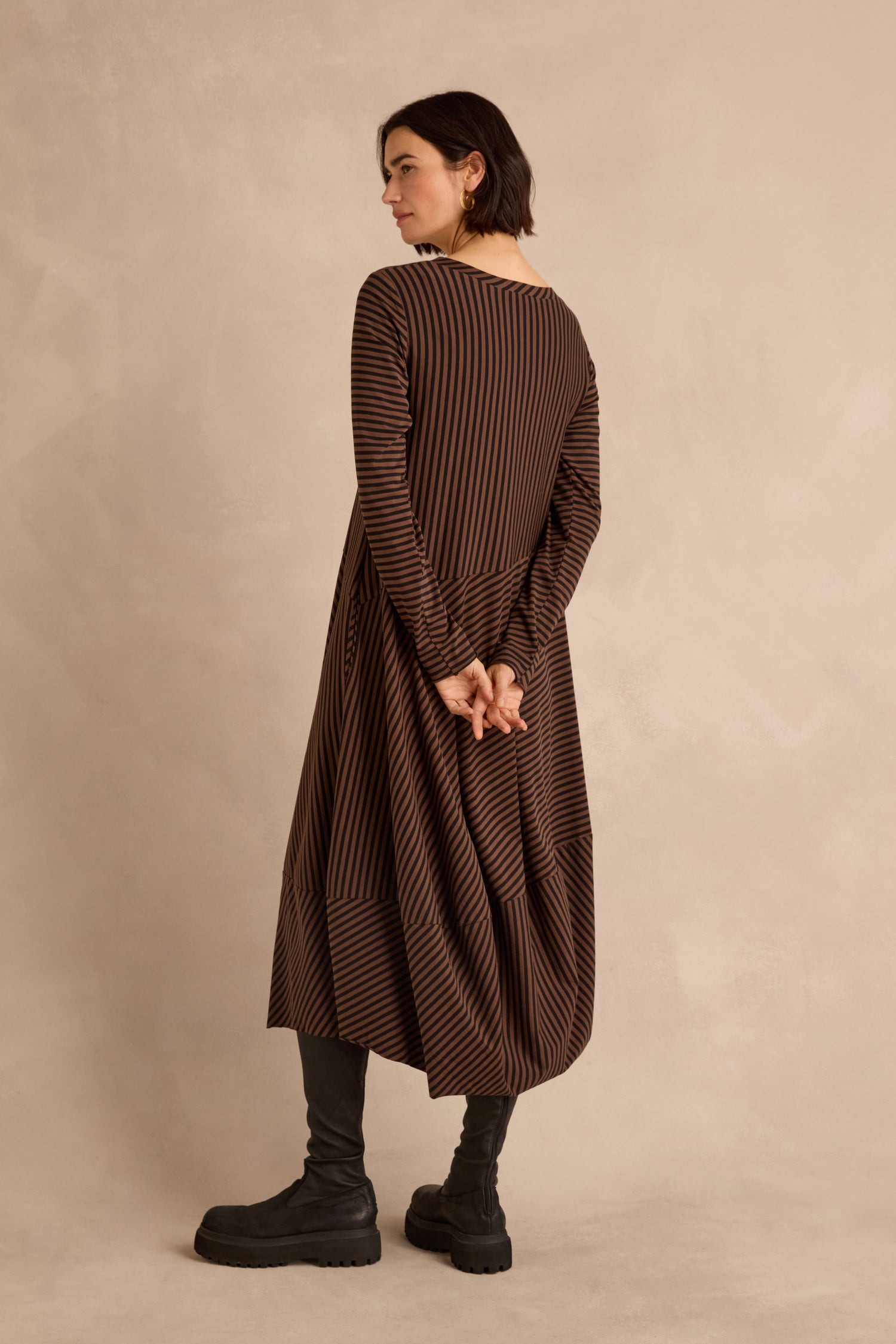 A person stands sideways with hands clasped behind their back, wearing the Draped Stripe Jersey Dress in brown and black stripes with black boots against a plain beige background.