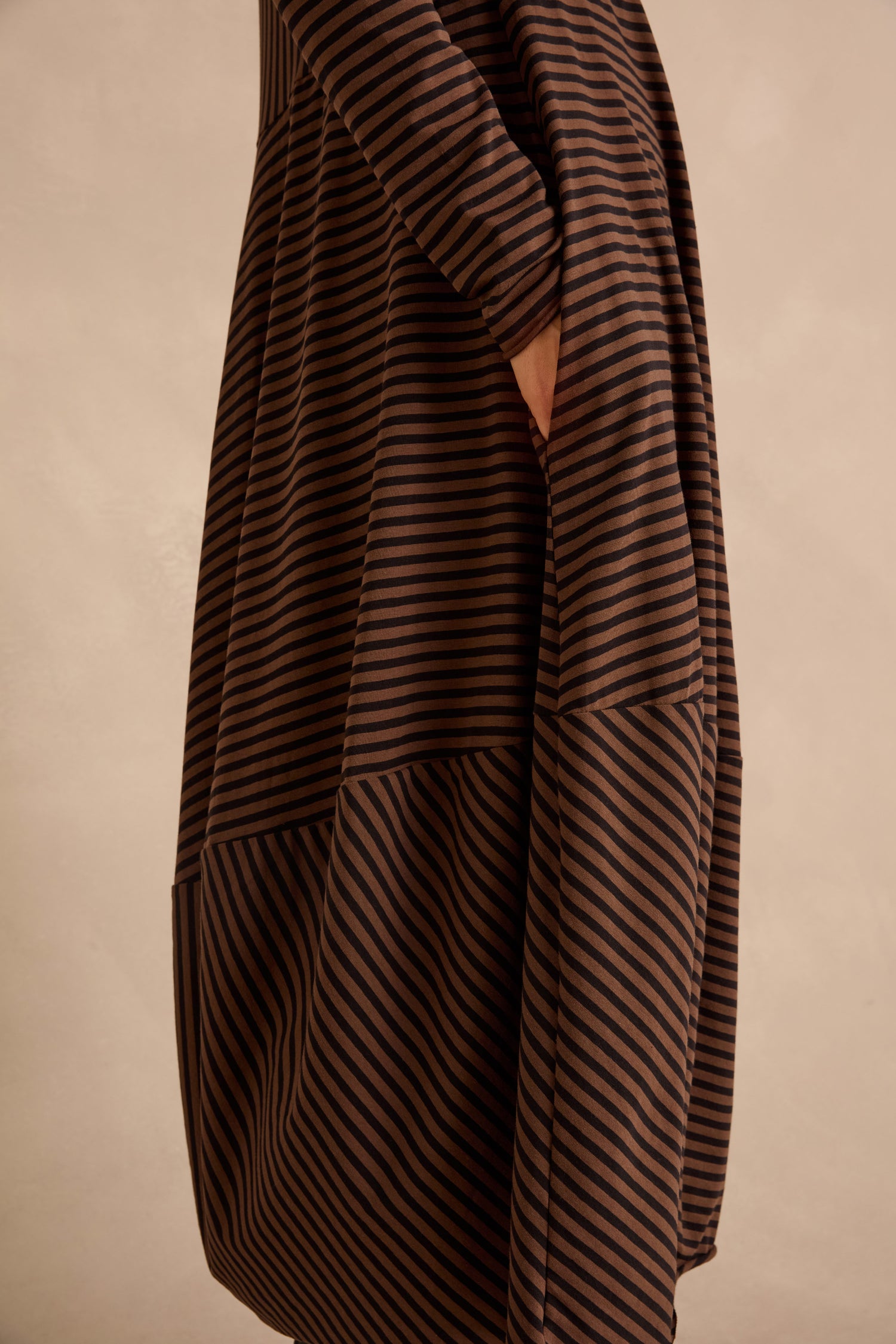 A person wearing the Draped Stripe Jersey Dress, featuring loose brown and black stripes and a side pocket, stands against a neutral background.