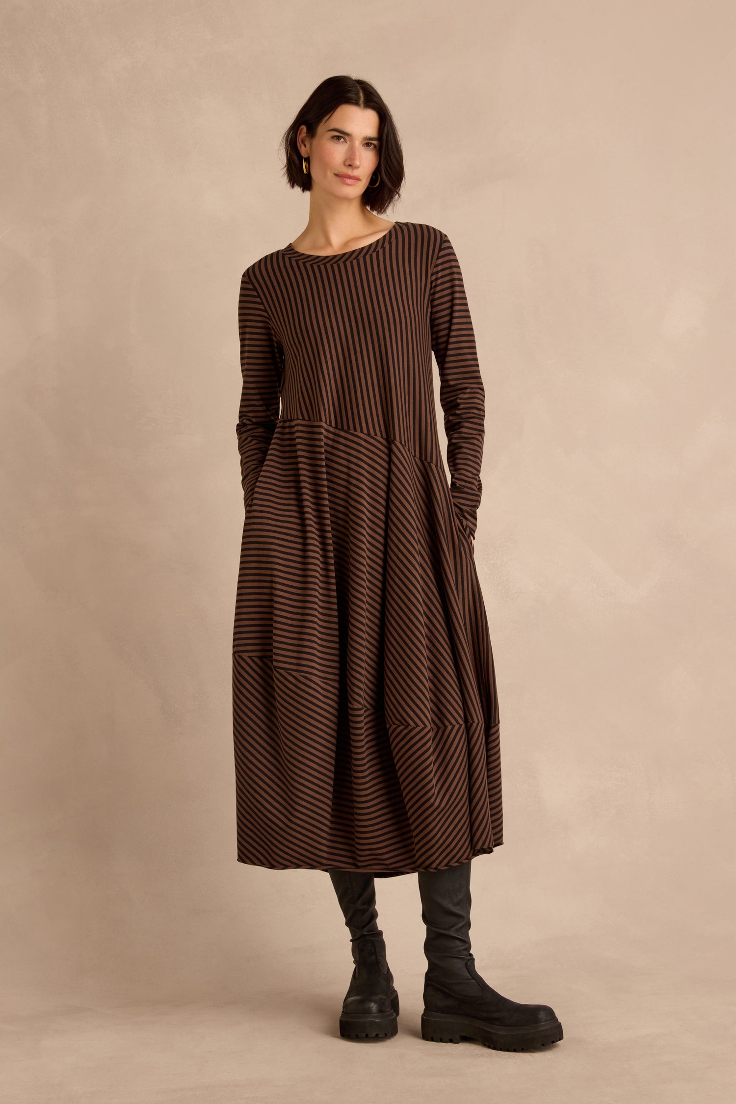 A woman stands against a beige background, wearing the Draped Stripe Jersey Dress— a long-sleeved brown and black striped style—paired with black leggings and black boots.