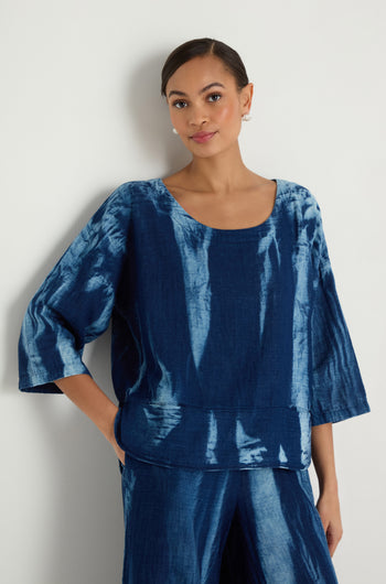 A woman stands against a white wall wearing the Soft Denim Two-Pocket Top, a loose-fitting blue tie-dye cotton denim piece with elbow-length sleeves, paired with matching blue pants.