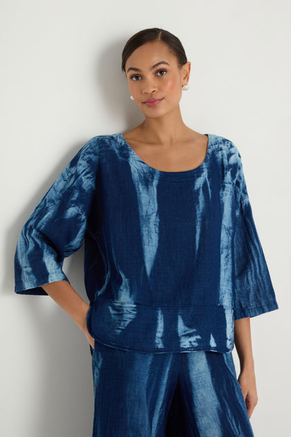 A woman stands against a white wall wearing the Soft Denim Two-Pocket Top, a loose-fitting blue tie-dye cotton denim piece with elbow-length sleeves, paired with matching blue pants.