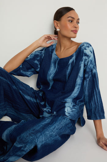 A woman models the Soft Denim Two-Pocket Top in blue tie-dye with matching pants, featuring a boxy silhouette. She sits against a white wall, gazing to the side with her hand resting near her neck.
