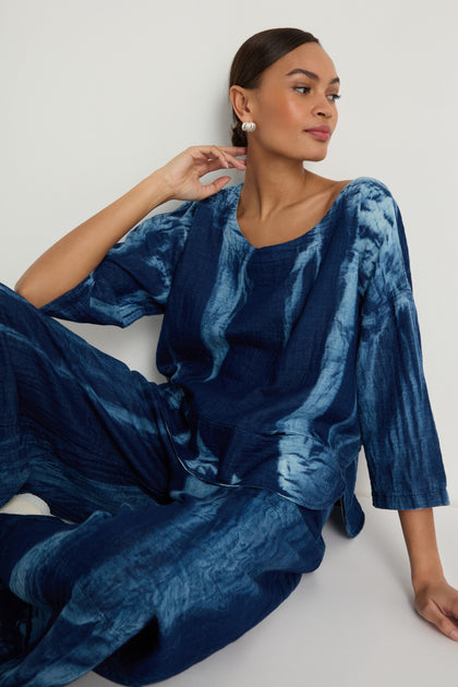 A woman models the Soft Denim Two-Pocket Top in blue tie-dye with matching pants, featuring a boxy silhouette. She sits against a white wall, gazing to the side with her hand resting near her neck.