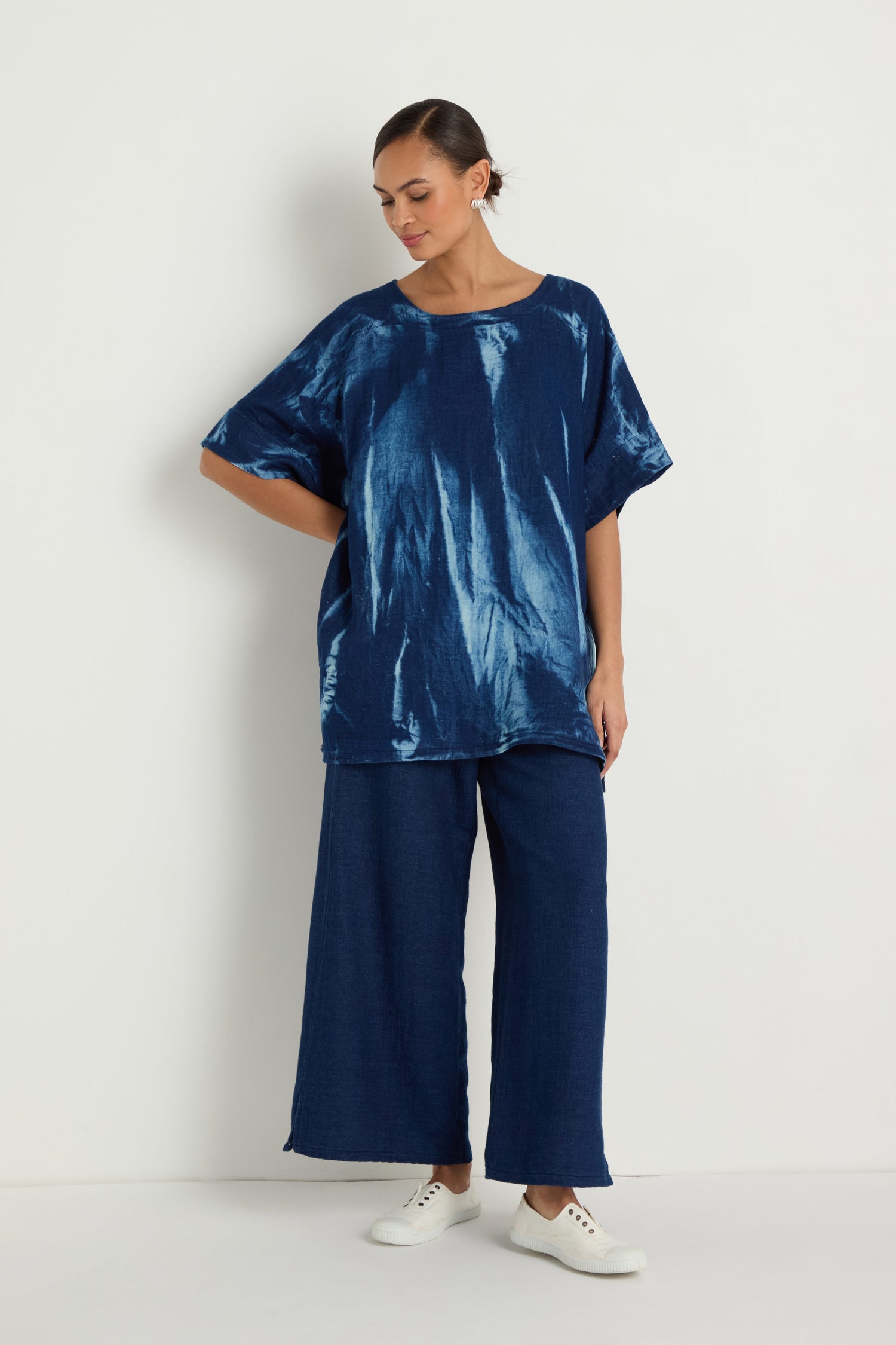 A woman stands against a white wall wearing the Soft Denim Button-Back Tunic with a longline silhouette, paired with wide-leg dark blue pants and white sneakers. She looks down, one hand resting on her hip.