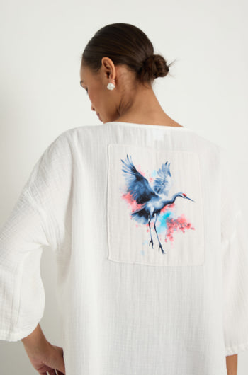 A person wears the Crinkle Two-Pocket Top in white, featuring a colorful crane illustration on the back, standing against a plain light background.