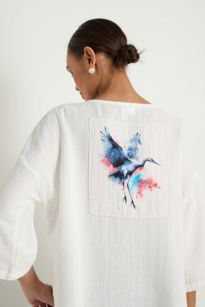 A person wears the Crinkle Two-Pocket Top in white, featuring a colorful crane illustration on the back, standing against a plain light background.