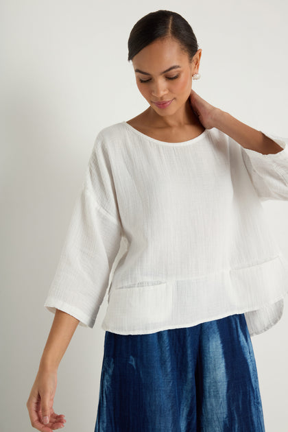 A woman wearing the Crinkle Two-Pocket Top in white and a blue skirt stands against a plain light background, looking down with one hand raised near her head.