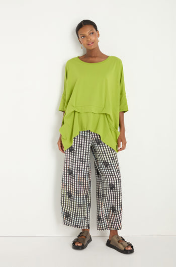 A woman stands against a white wall wearing the Tuck Hem Swing Lime Tunic in a cotton blend, paired with black and white checkered wide-leg patterned pants and black sandals.
