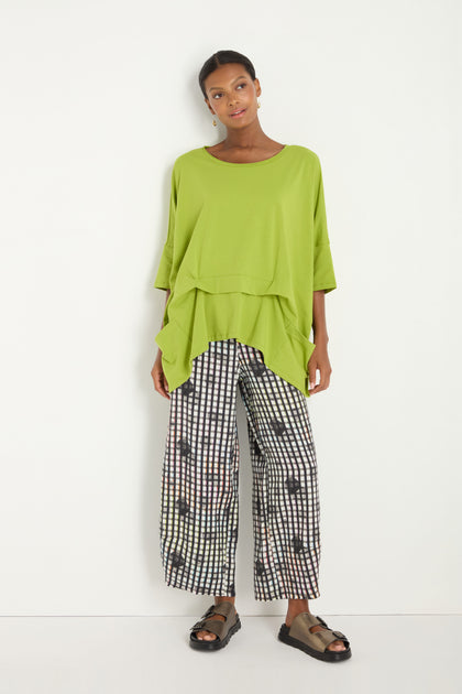 A woman stands against a white wall wearing the Tuck Hem Swing Lime Tunic in a cotton blend, paired with black and white checkered wide-leg patterned pants and black sandals.