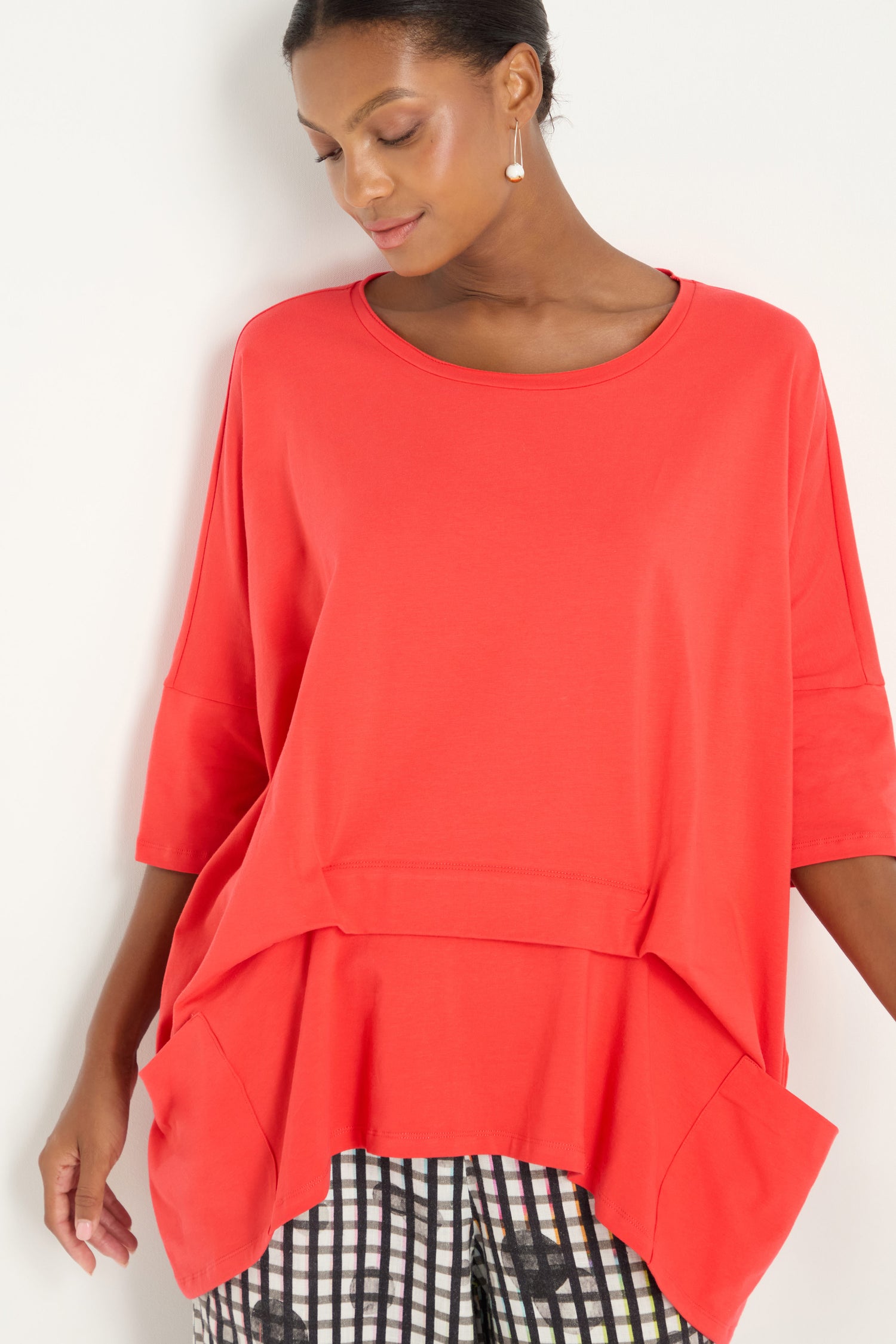 A woman models the Tuck Hem Swing Red Tunic with elbow-length sleeves, paired with patterned black-and-white pants, against a plain white background.
