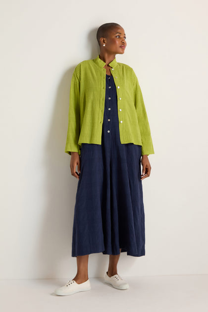 A person wears a navy blue, ankle-length dress paired with the Self Check Lime Boxy Shirt and white sneakers, standing against a plain light-colored wall.