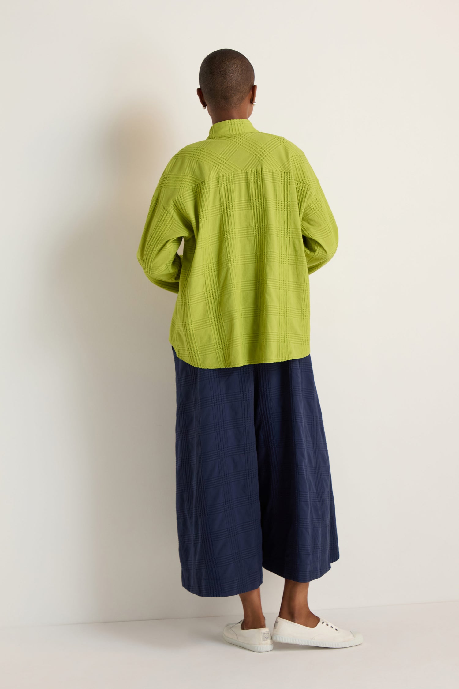 A person with a shaved head is seen from behind, wearing the Self Check Lime Boxy Shirt, wide navy blue pants, and white sneakers against a plain light background, highlighting the shirt's relaxed silhouette.