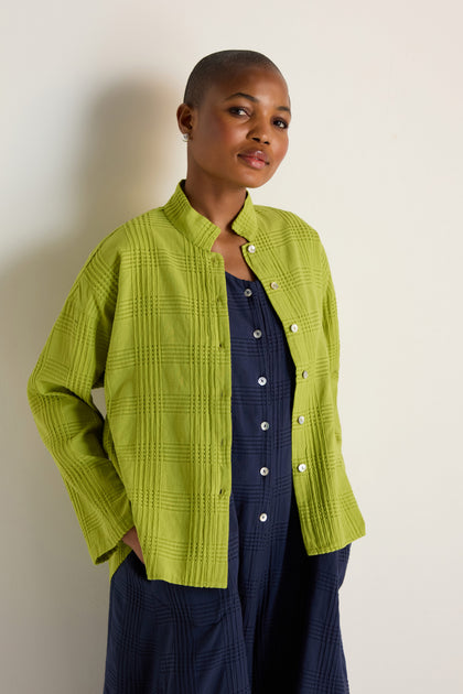 A person with short hair wears the Self Check Lime Boxy Shirt over a navy relaxed-fit buttoned dress, standing against a plain light-colored wall.