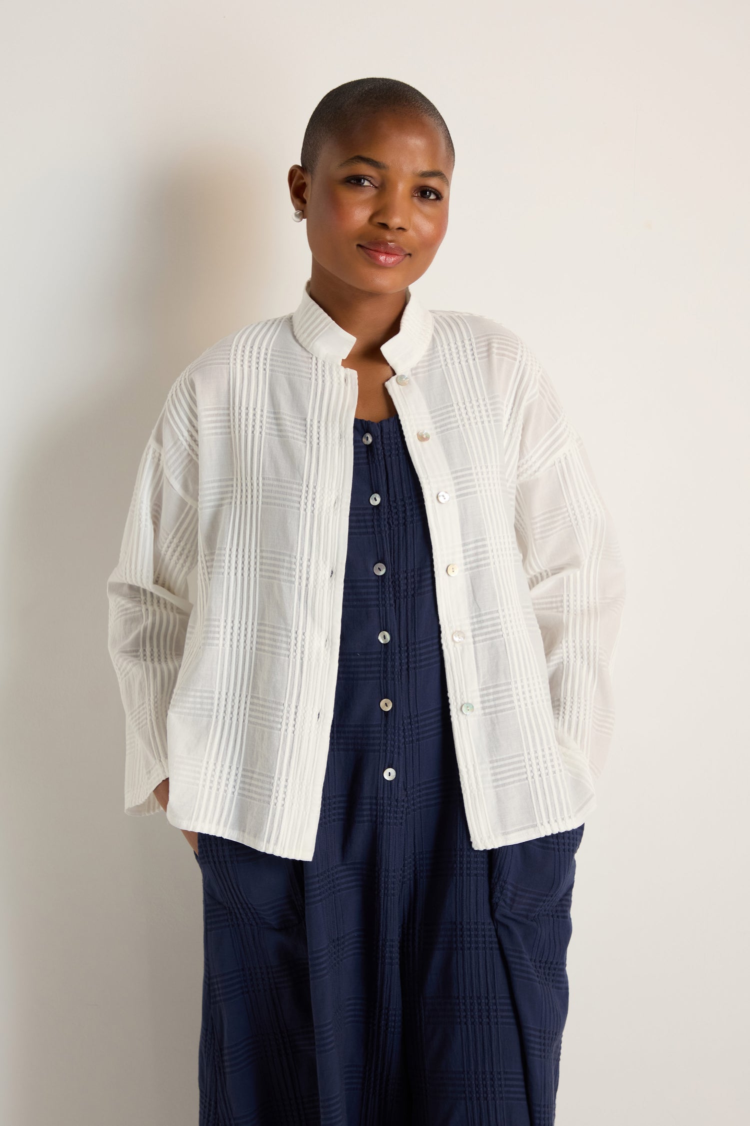 A person with short hair in a relaxed silhouette wears the Self Check White Boxy Shirt over a navy blue dress, standing against a plain light-colored wall.