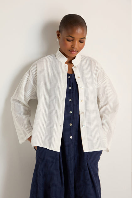 Wearing the Self Check White Boxy Shirt, featuring textured cotton and a relaxed silhouette, a person stands against a light wall with hands in pockets, looking down.