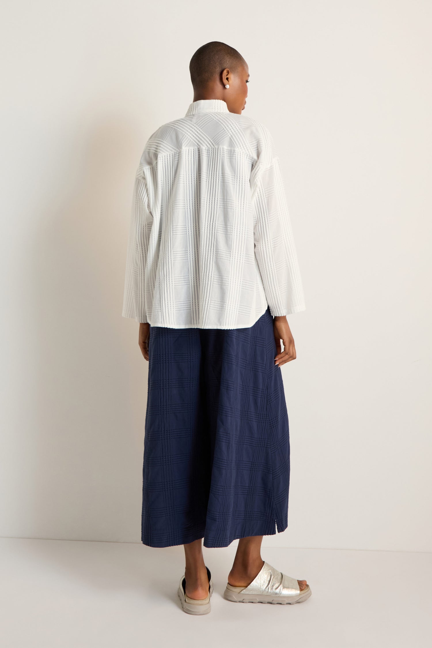 A person stands facing away, wearing the Self Check White Boxy Shirt with a relaxed fit, wide navy pants, and light sandals against a plain white background.