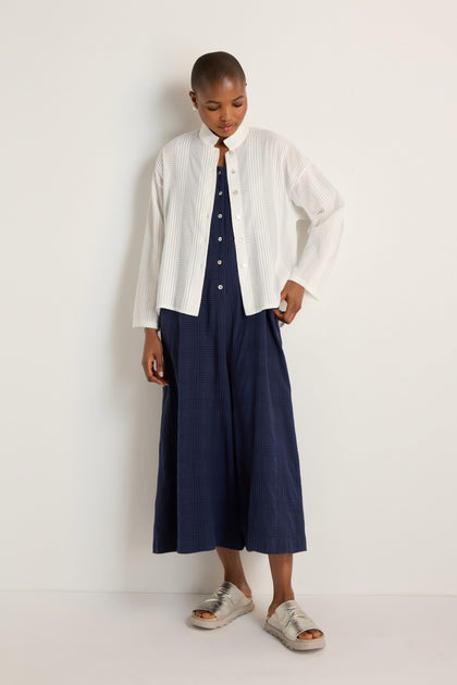 Person stands against a white wall, wearing the Self Check White Boxy Shirt in a relaxed boxy silhouette over a navy blue dress and paired with silver sandals.