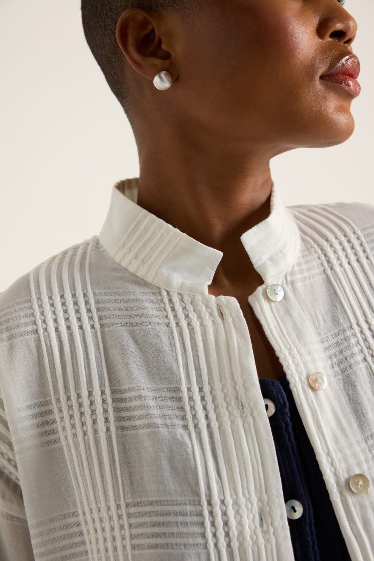 A person wears the Self Check White Boxy Shirt in white cotton with vertical pleats and iridescent buttons, featuring a relaxed fit. Pearl stud earrings complete the look.