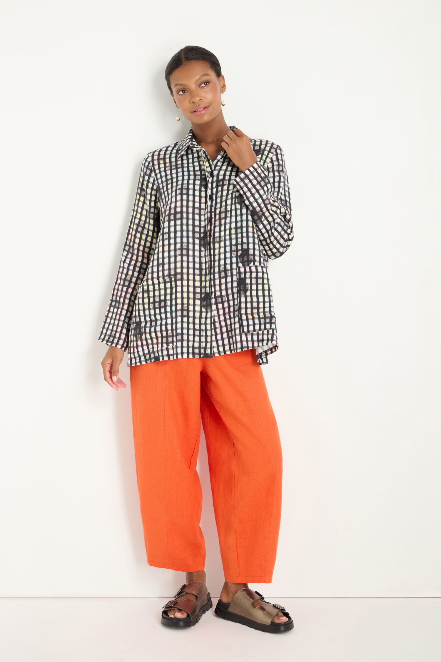 A person stands against a white background wearing a relaxed fit Check Dot Linen Shirt, orange wide-leg pants, and black sandals.
