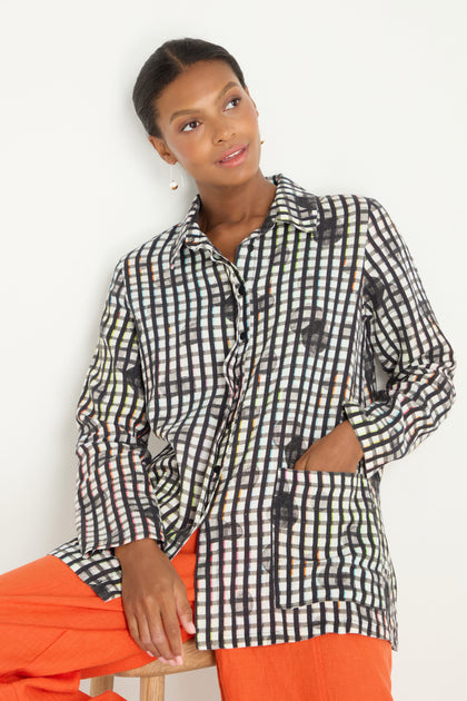 A woman sits against a white background, wearing orange pants and the Check Dot Linen Shirt—a relaxed fit, long-sleeve, black and white check dot top—with one hand in her pocket.