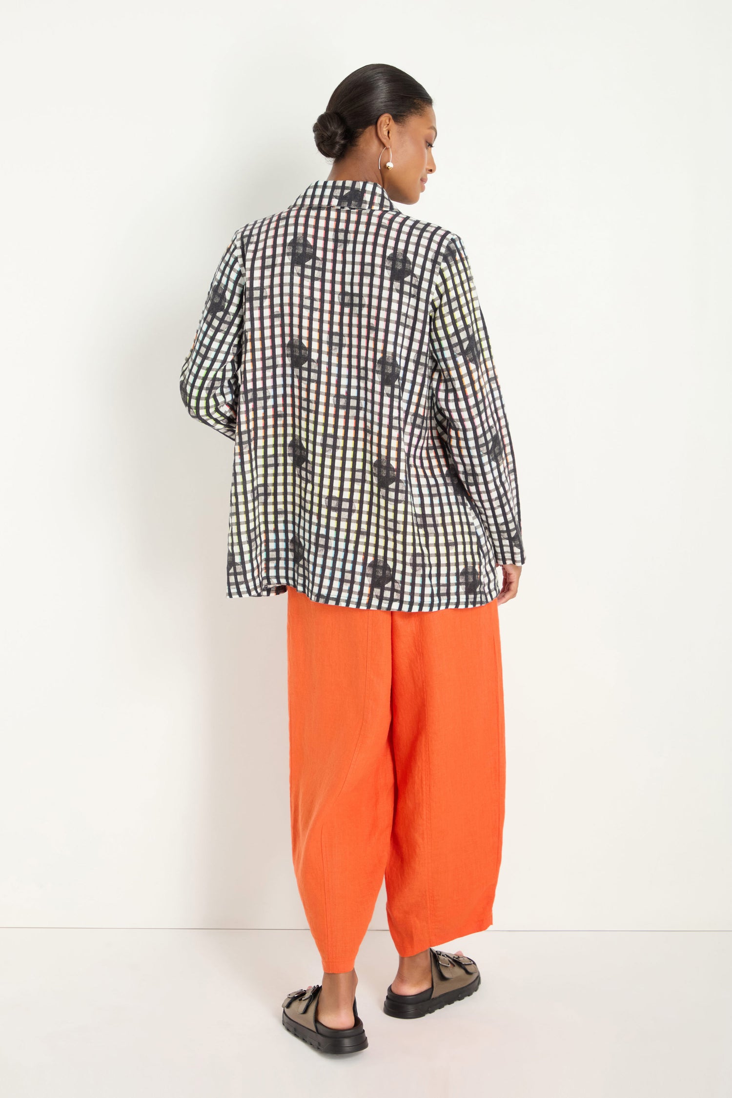 A person stands against a white background wearing the Check Dot Linen Shirt, featuring a relaxed fit with black-and-white abstract checkered patterns, paired with bright orange loose-fitting pants and black sandals.