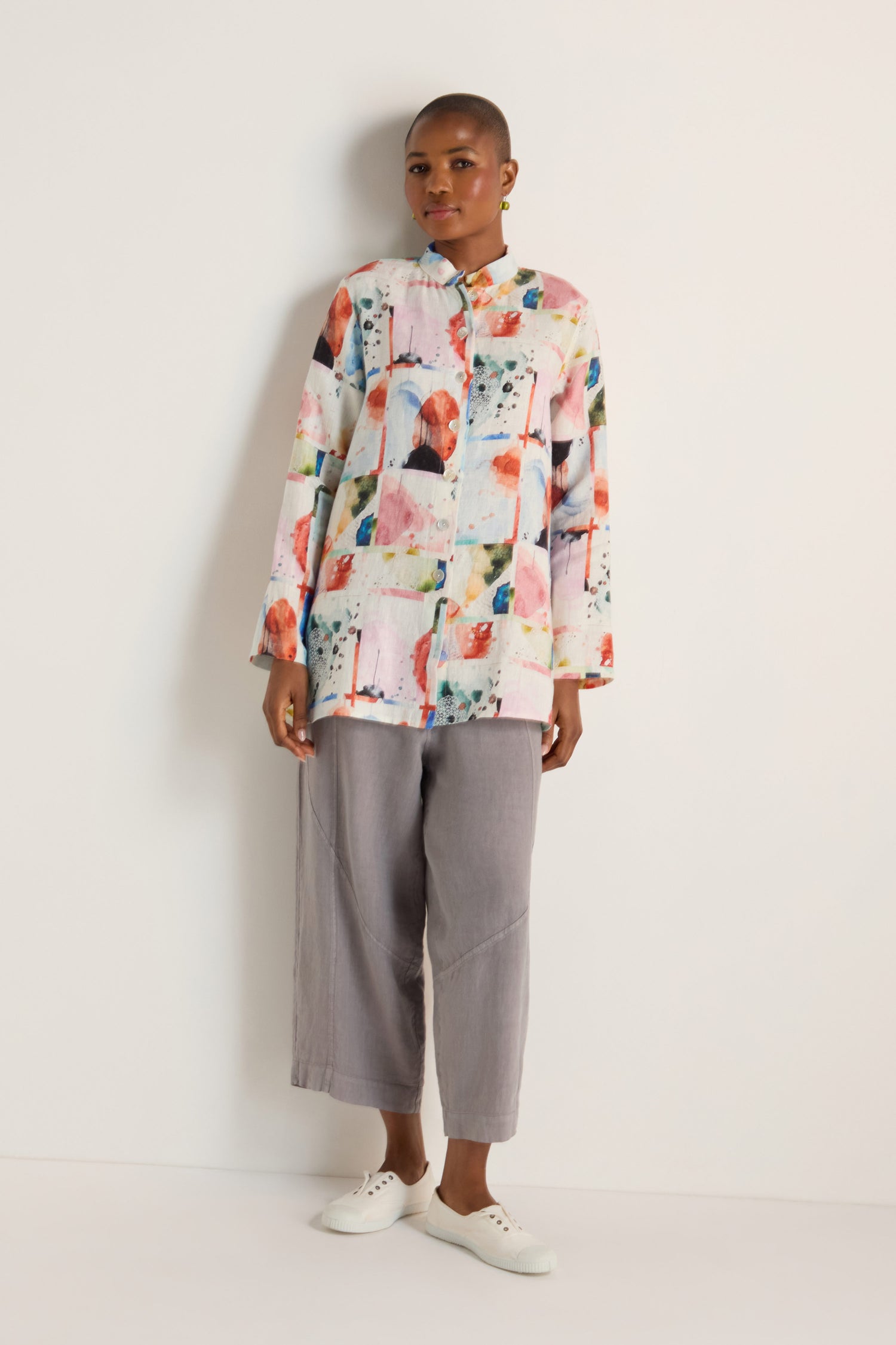 A person stands against a white wall, wearing a relaxed fit Watercolour Linen Print Shirt with long sleeves, paired with loose gray pants and white slip-on shoes.