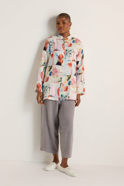 A person stands against a white wall, wearing a relaxed fit Watercolour Linen Print Shirt with long sleeves, paired with loose gray pants and white slip-on shoes.