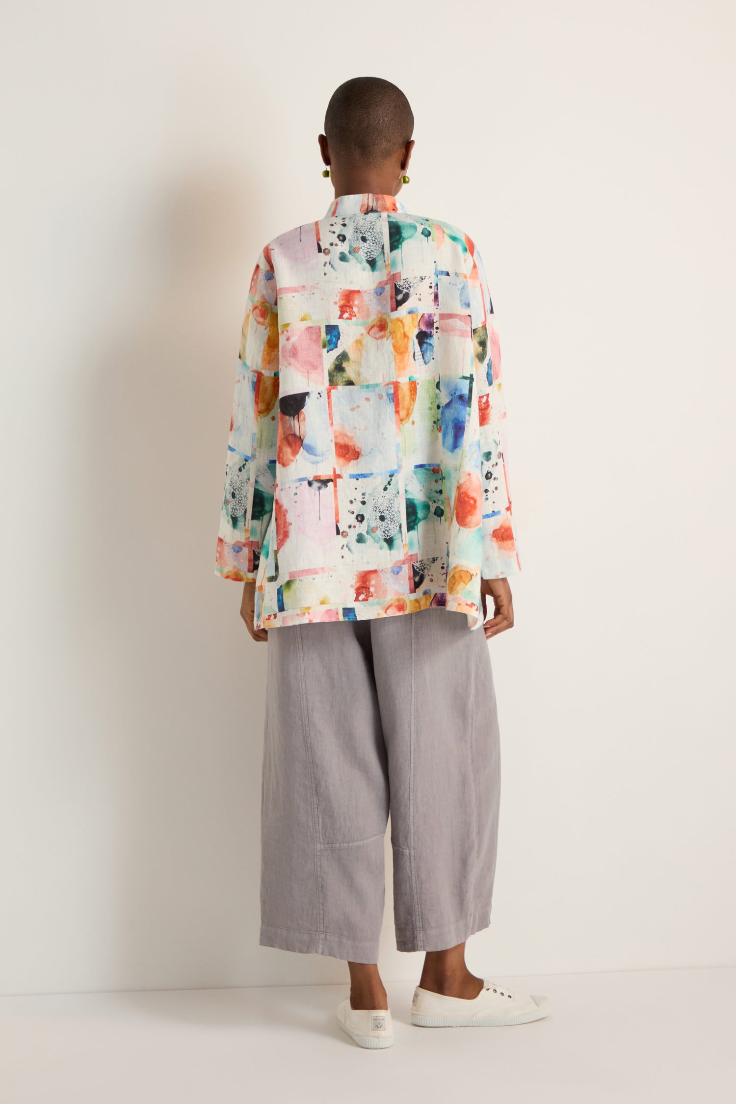 A person stands against a plain background, seen from the back, wearing the Watercolour Linen Print Shirt with an abstract pattern, loose gray pants, and white slip-on shoes.