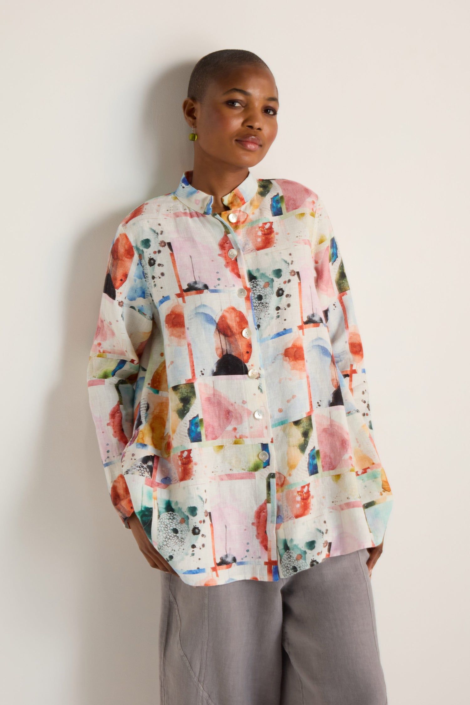 A person stands against a plain wall, wearing the Watercolour Linen Print Shirt with a colorful abstract pattern and light gray pants.